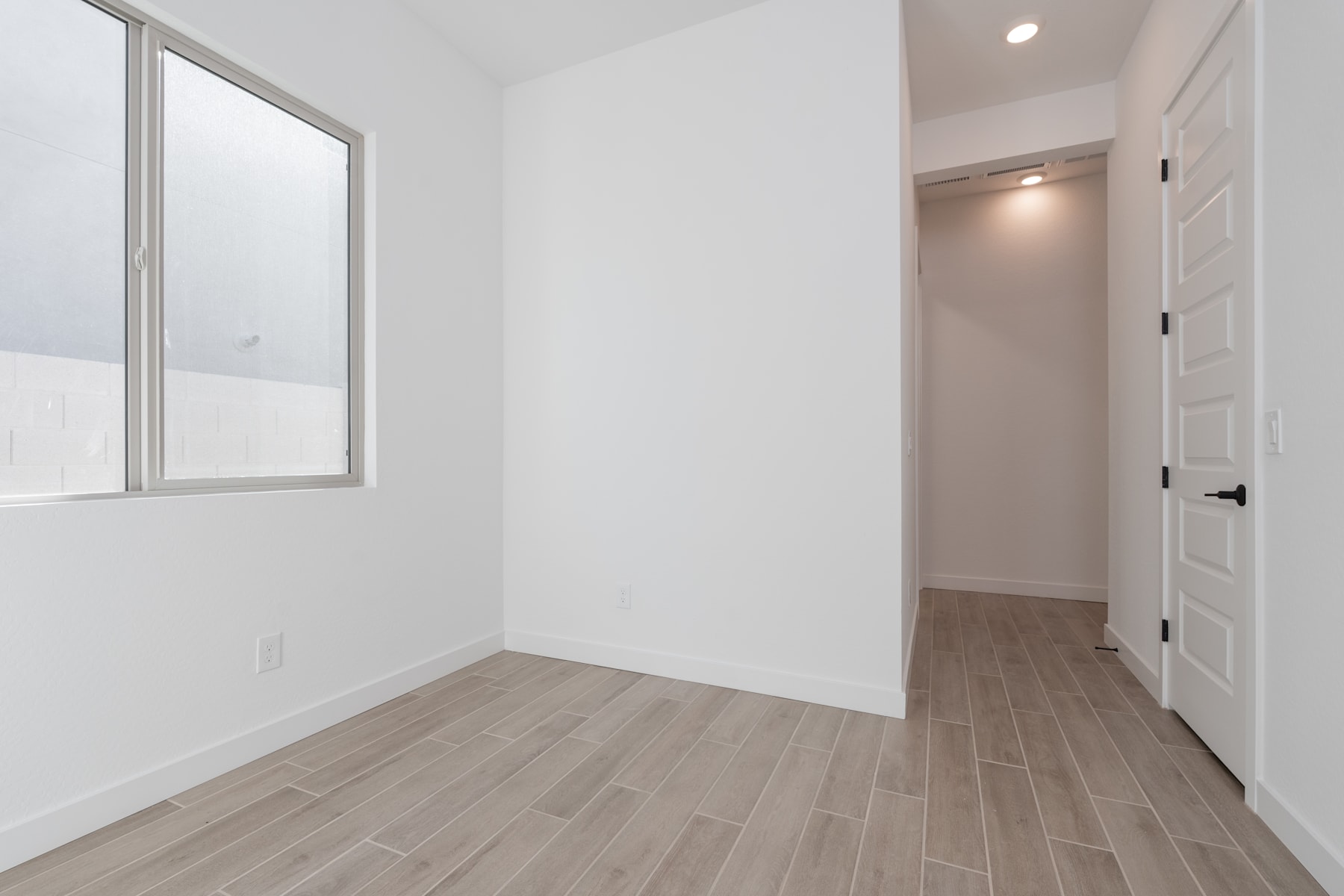 A bright, minimalist room with a large window, hardwood-style flooring, and a doorway leading to another area.
