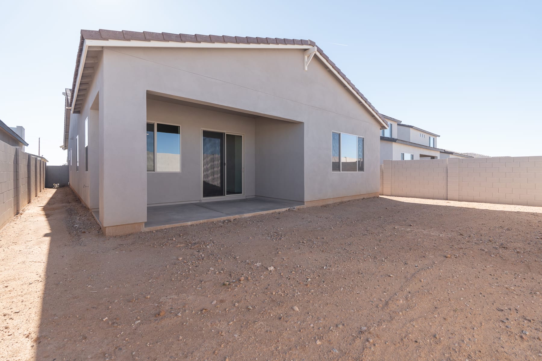 A single-story, beige-colored residential building with a slanted roof stands in the foreground, surrounded by a dirt-covered yard and a fence in the background.