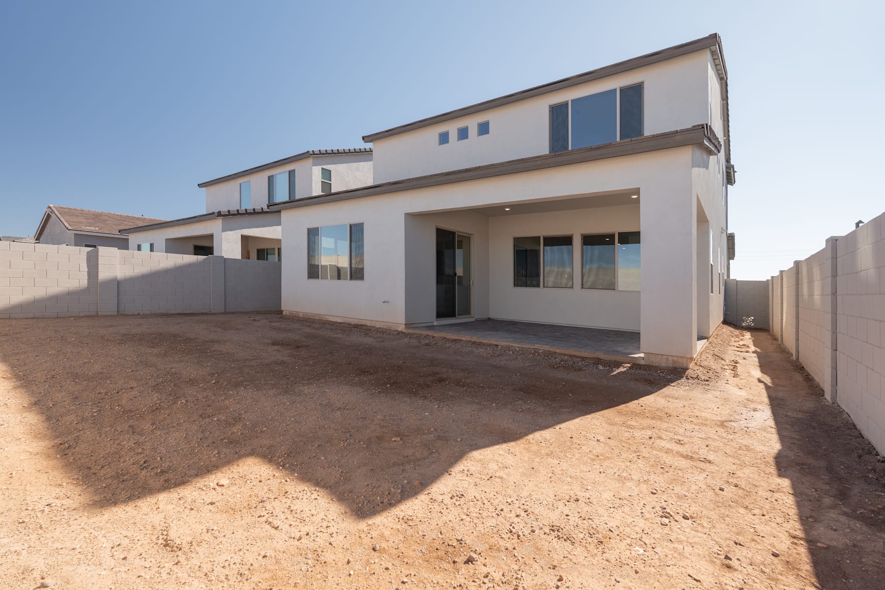 A modern two-story residential building with a paved driveway and a dirt-covered yard in the foreground, set against a clear blue sky.