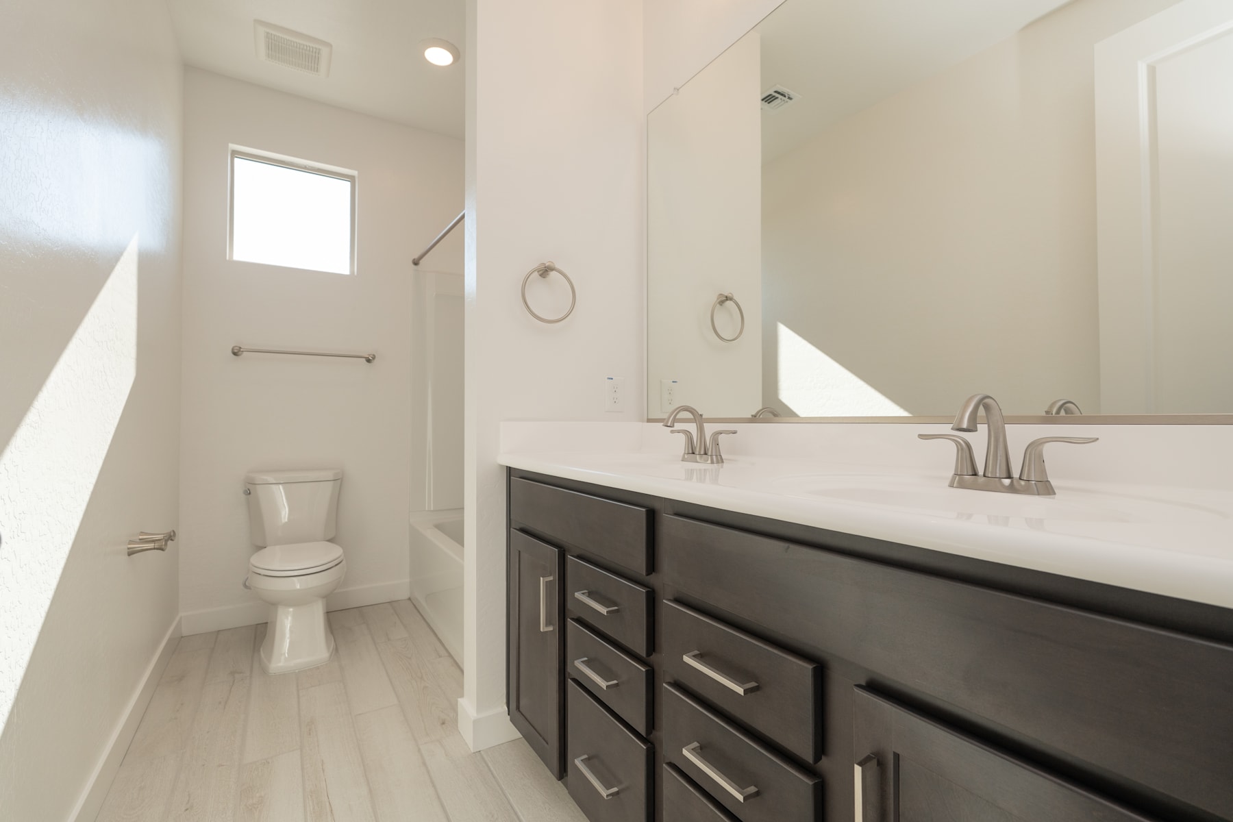 A modern bathroom with a dark vanity cabinet, white countertop, and a toilet in the background, all set against a bright and airy environment.