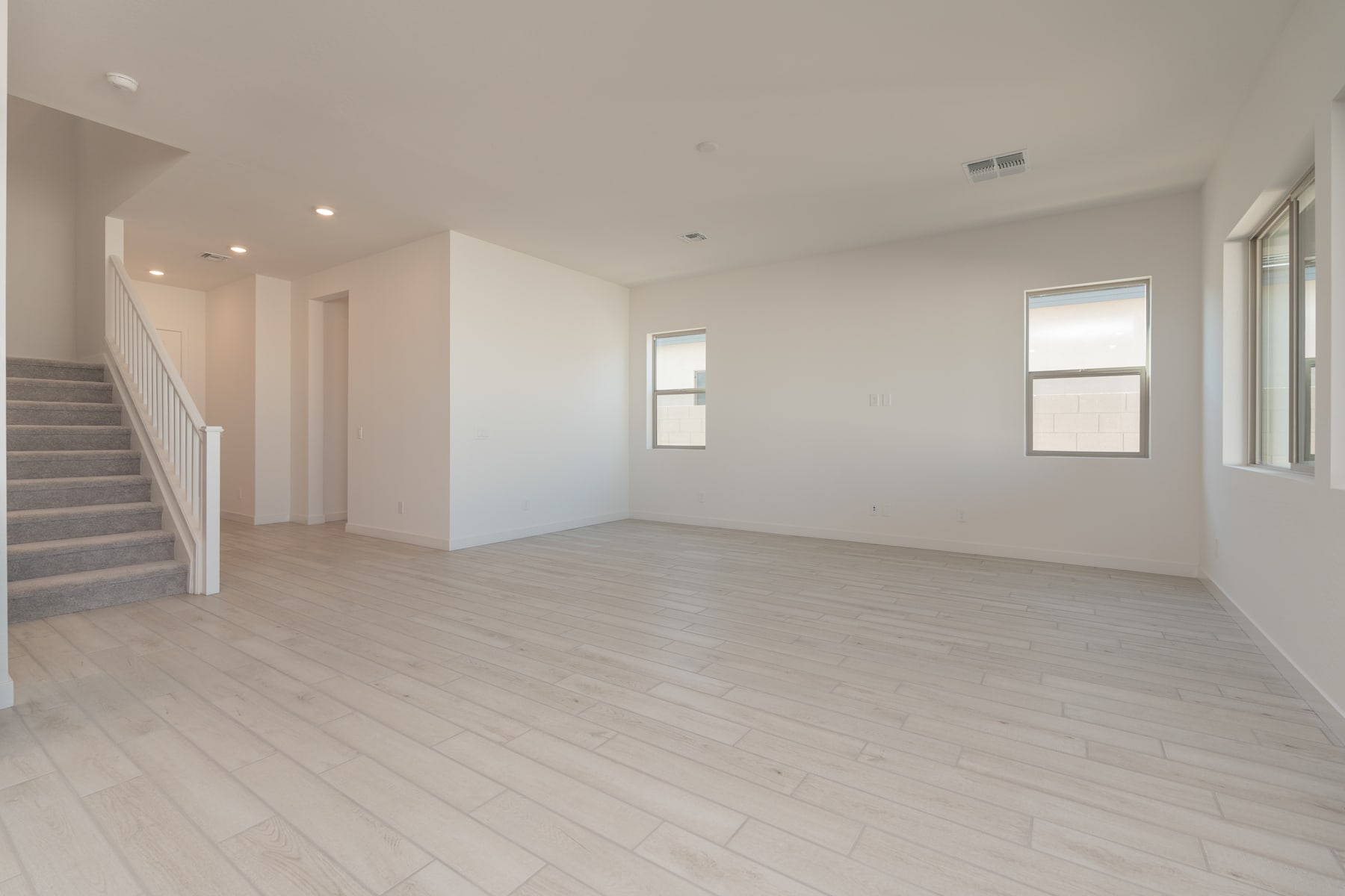 A spacious, minimalist room with light-colored hardwood floors, white walls, and a staircase visible in the background.