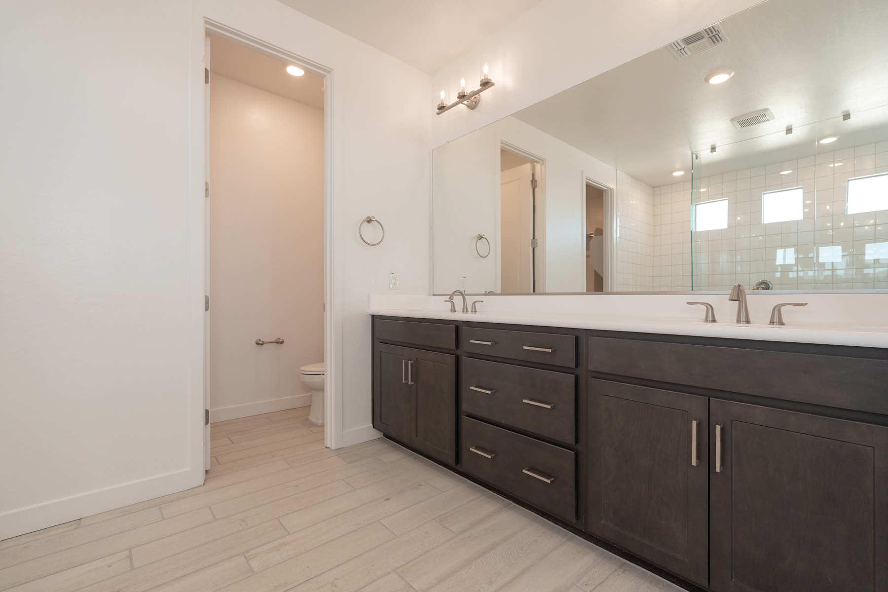 A modern and spacious bathroom with a large vanity, double sinks, and a well-lit mirror. The floor is covered in light-colored wood-like tiles, creating a clean and minimalist aesthetic.
