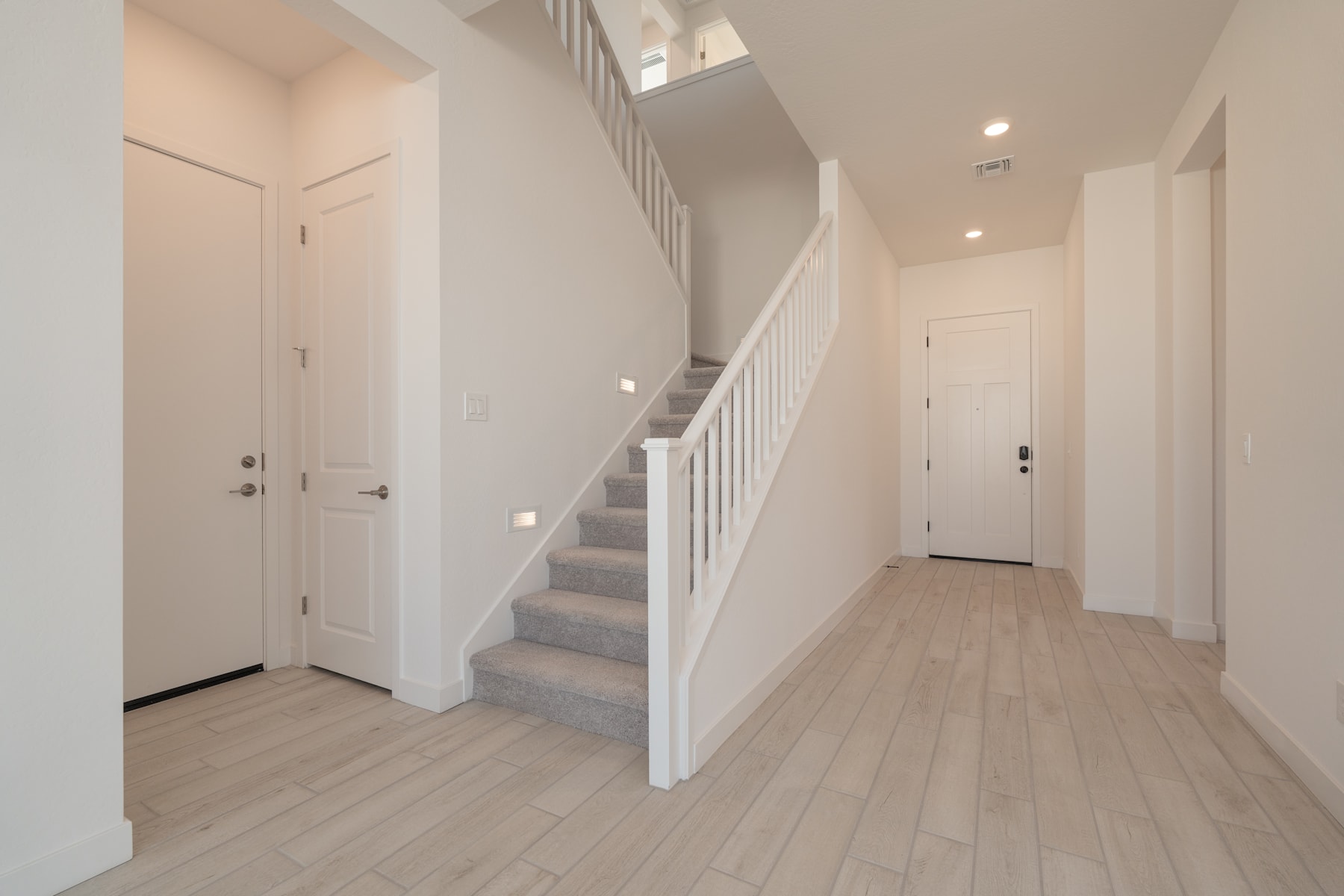 A bright and spacious hallway with a staircase leading upstairs, featuring white walls, a wooden floor, and recessed lighting.