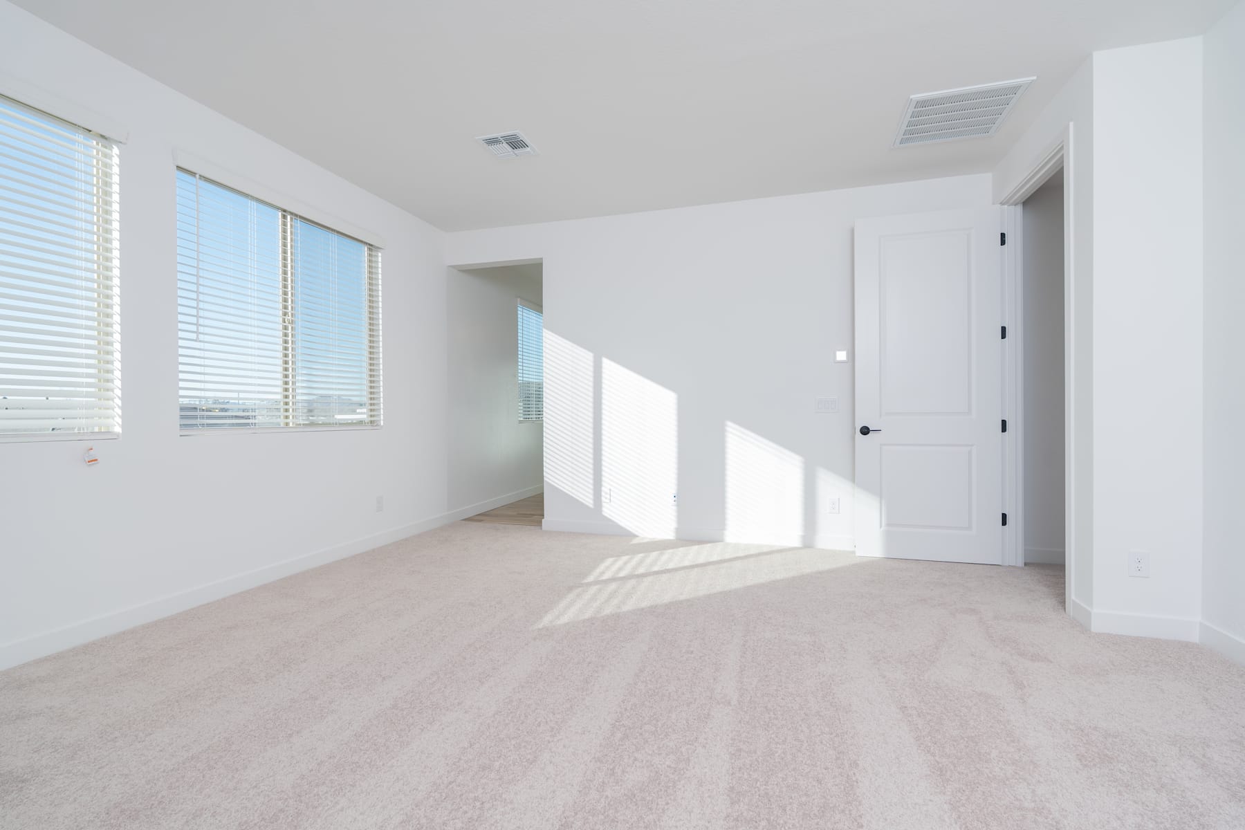 A bright, spacious room with large windows, white walls, and a light-colored hardwood floor. The room appears to be empty, creating a sense of openness and minimalism.