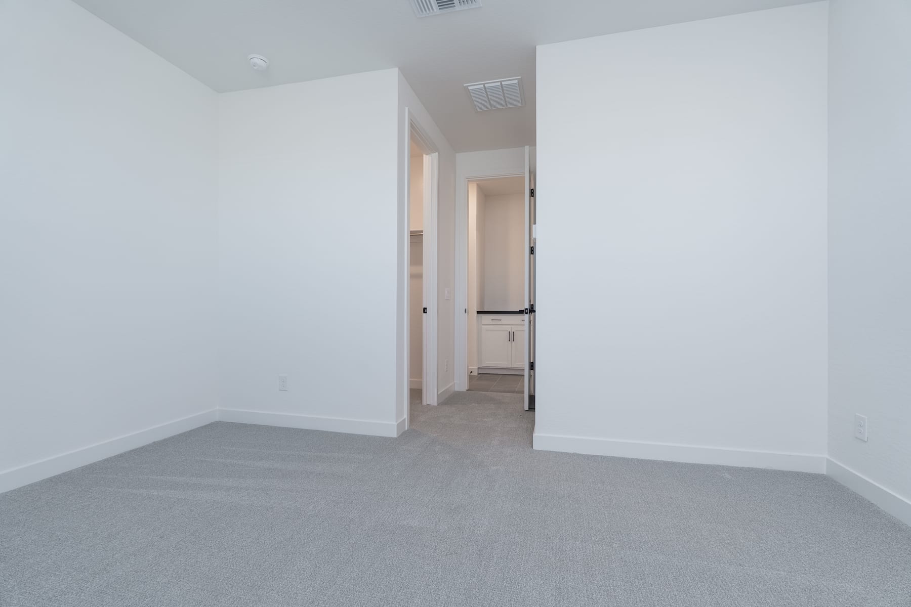 A spacious, minimalist room with white walls and a gray carpeted floor, leading to a doorway in the background.