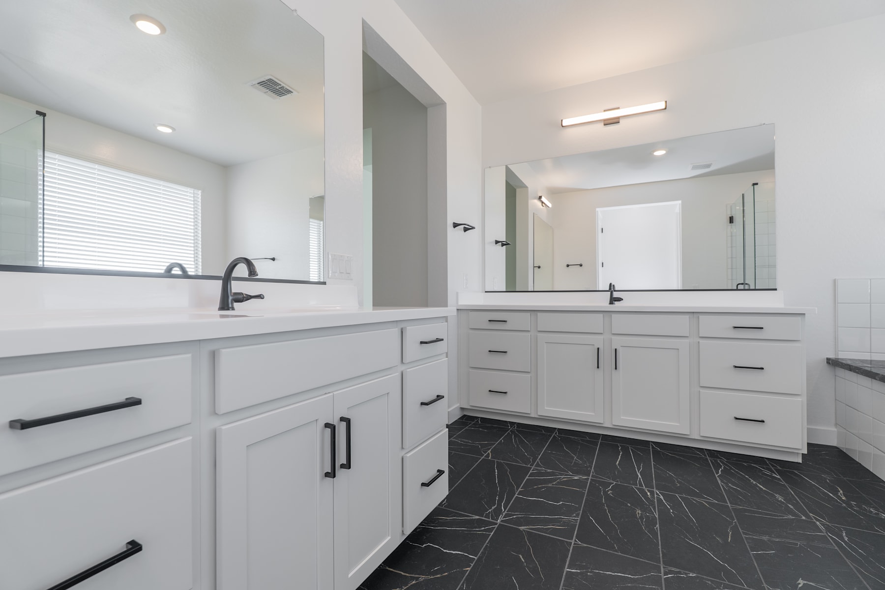 A modern, well-lit bathroom with white cabinets, a double vanity, and a tiled floor in a dark, marble-like pattern.