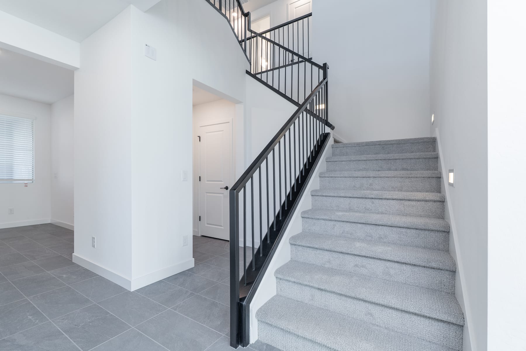 A modern, minimalist staircase with gray tile steps and a black metal railing leads up to a doorway in a bright, open space.
