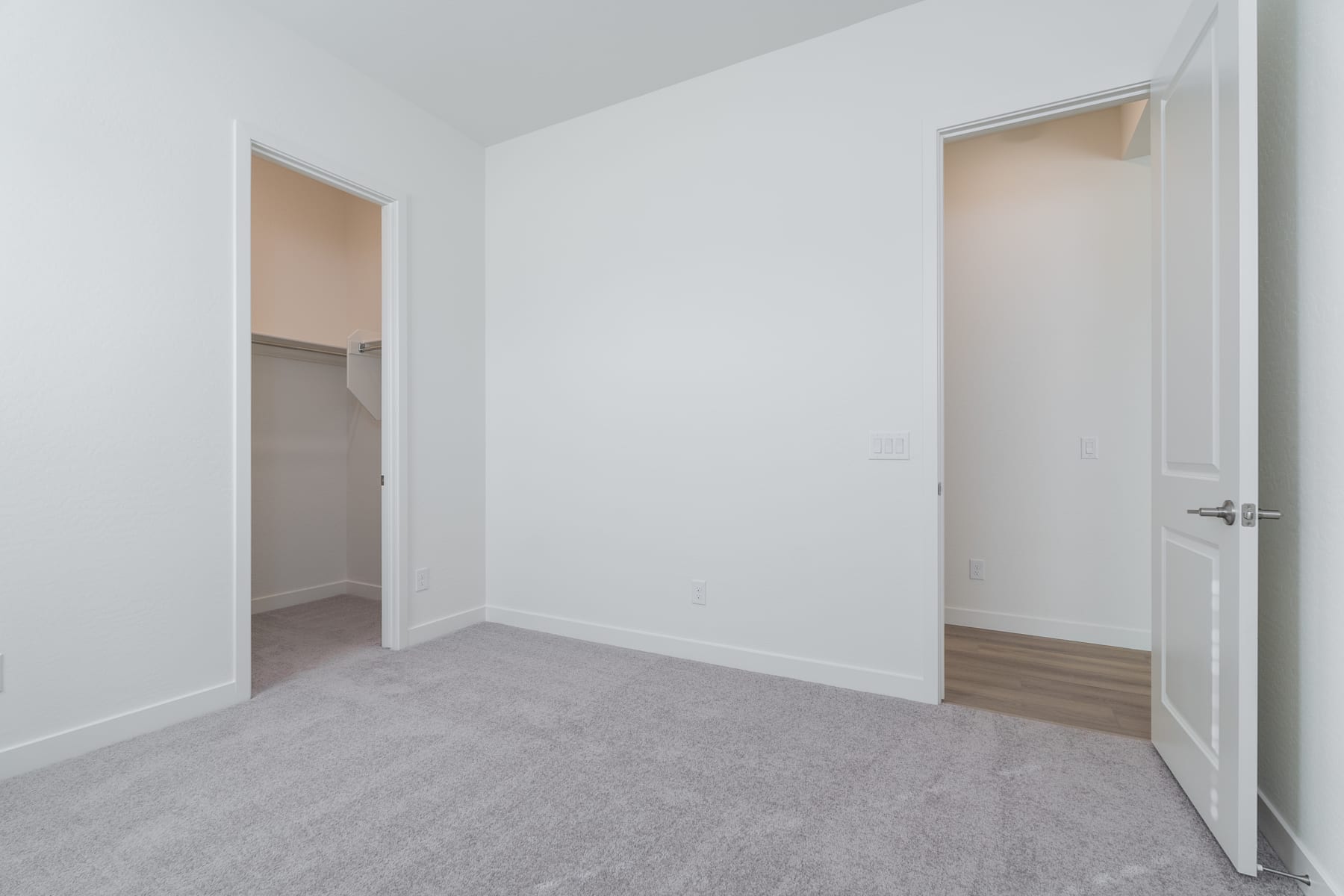 A spacious, minimalist room with white walls, a gray carpeted floor, and an open doorway leading to another room.