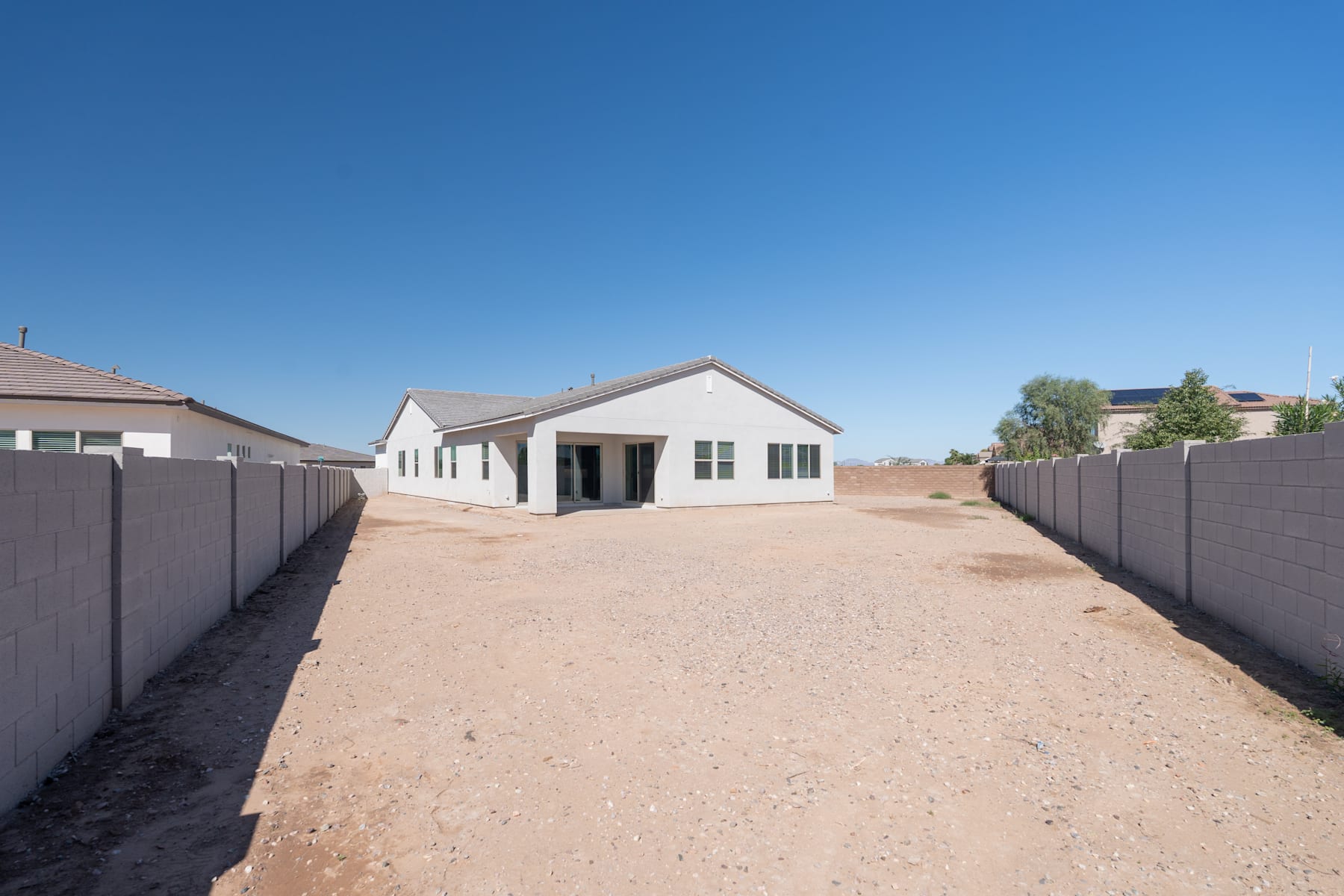A white, single-story house with a gravel driveway leading up to it, surrounded by a clear blue sky and a fenced-in yard in the foreground.