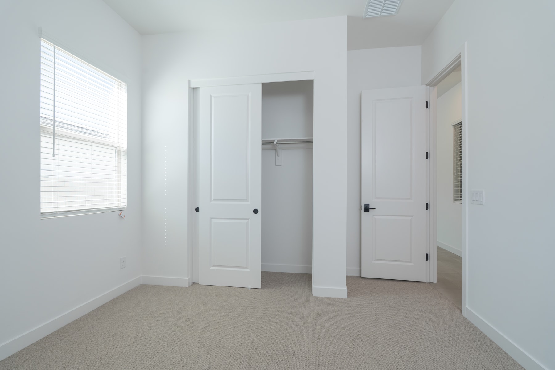 A bright, minimalist bedroom with white walls, a window, and two closet doors in the foreground.