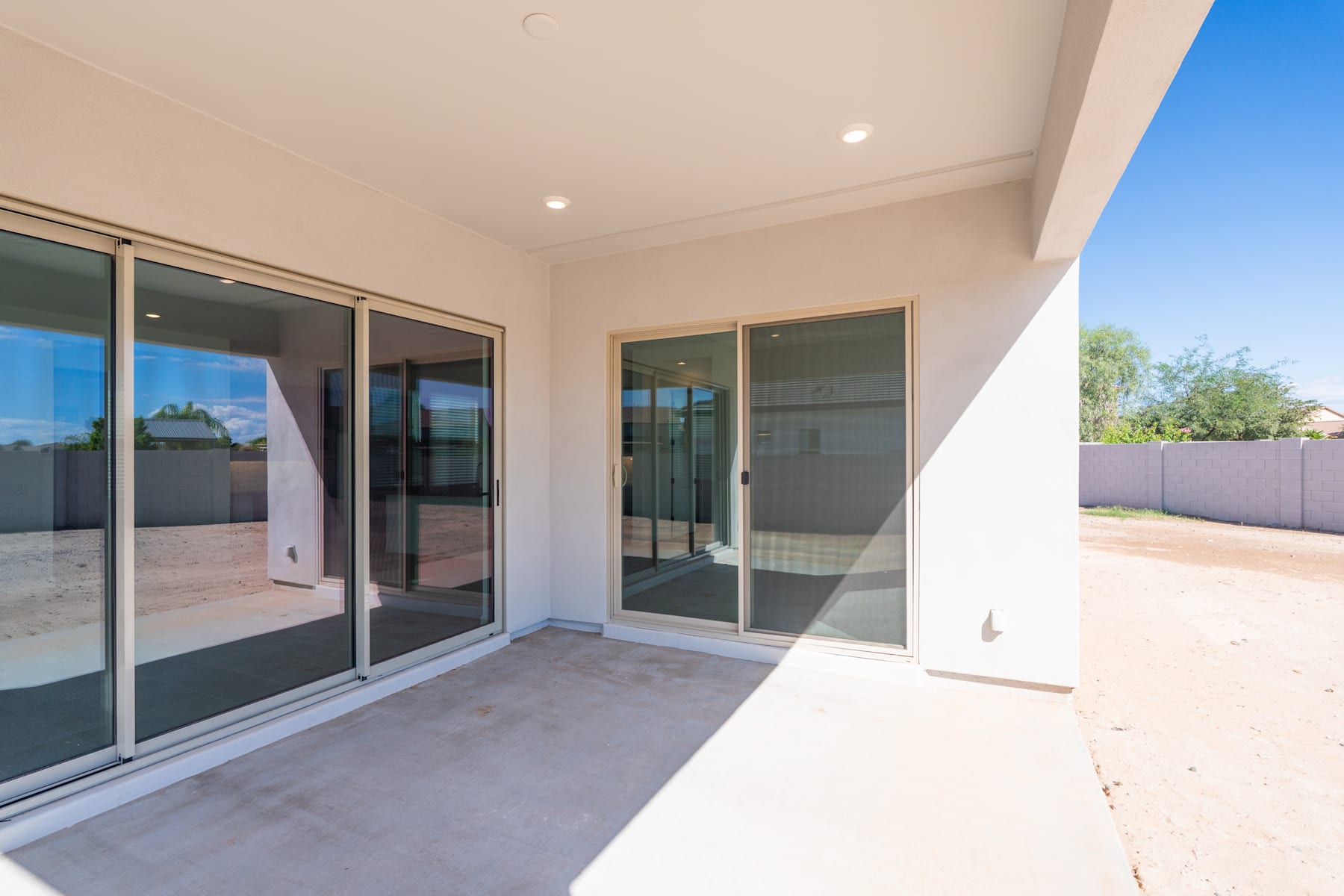 The image shows a modern, open-plan patio or outdoor living area with large sliding glass doors leading to the interior of the home. The patio has a concrete floor and the background features a clear blue sky, suggesting a warm, sunny climate.