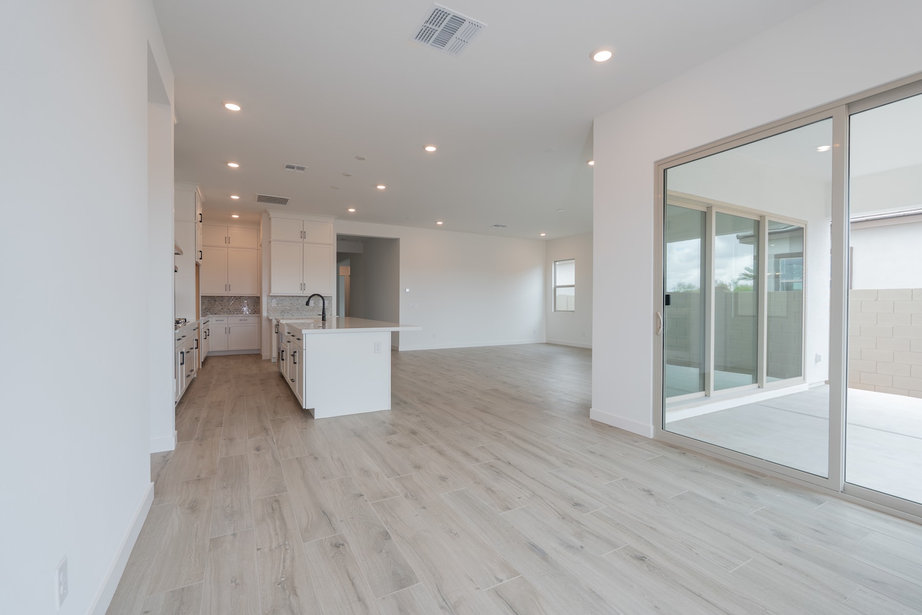 A spacious, open-concept living area with a kitchen visible in the background, featuring light-colored hardwood flooring and large windows that allow natural light to flood the space.