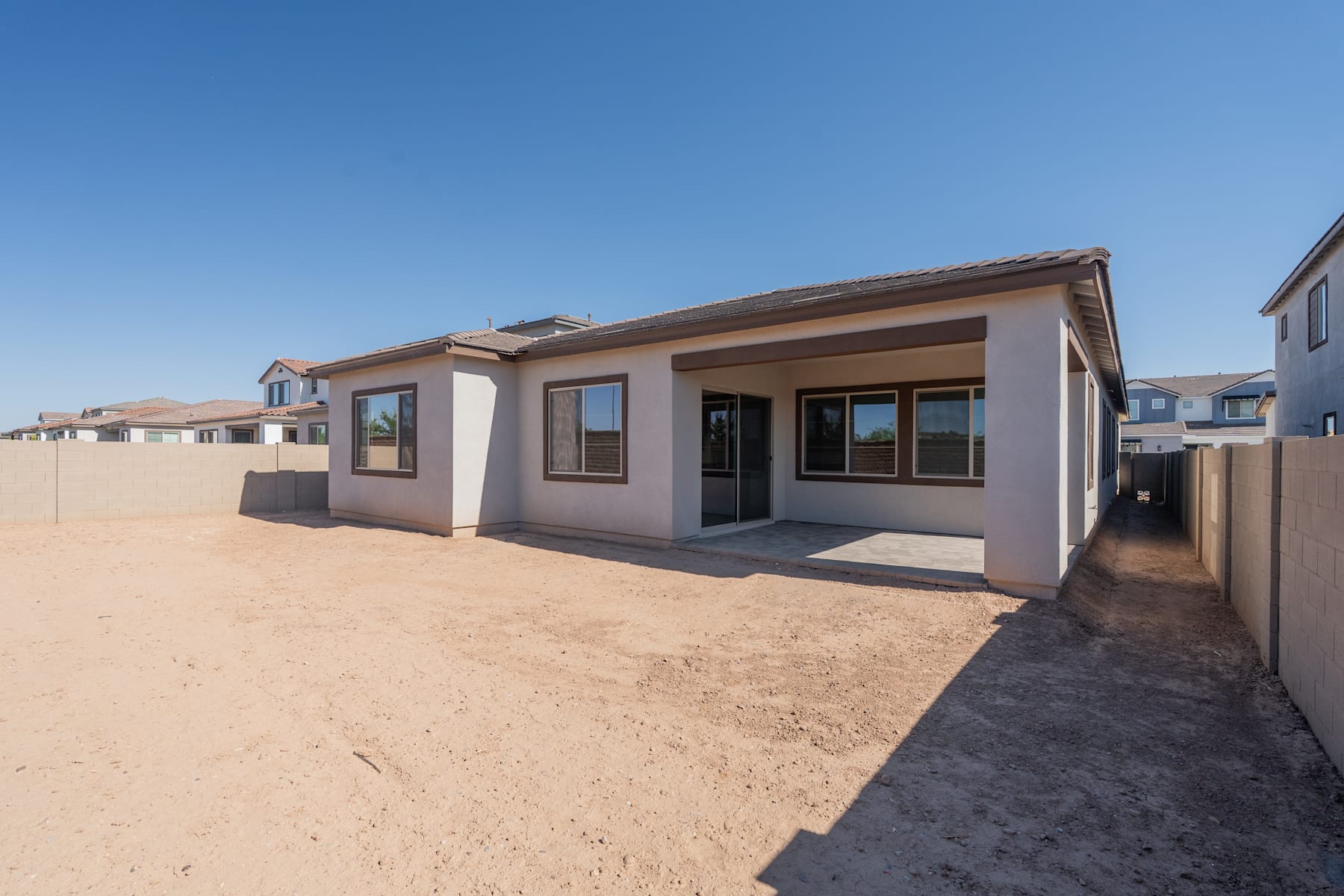 A single-story residential building with a beige exterior and a tiled roof stands in the foreground, surrounded by a sandy landscape and clear blue sky in the background.