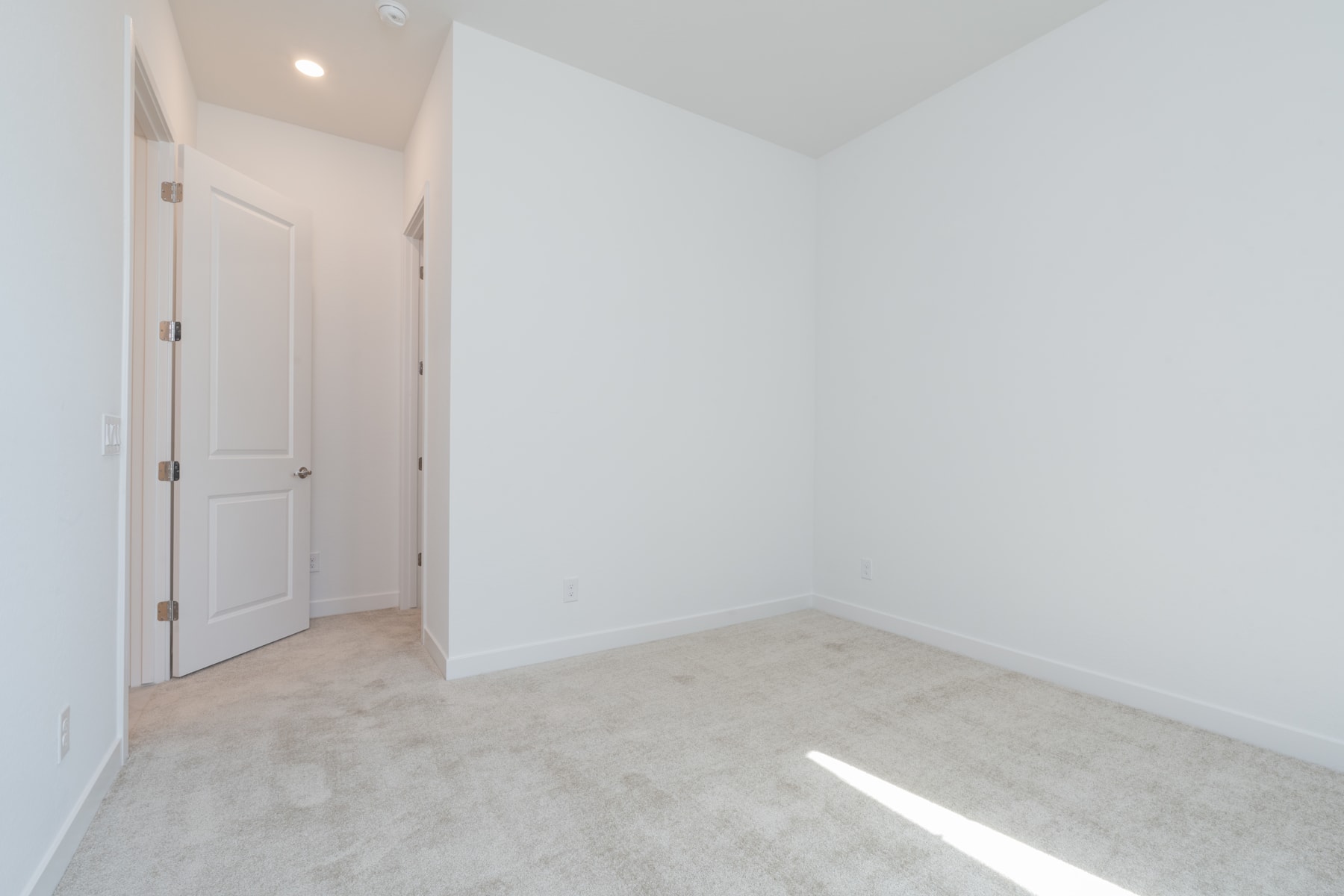 A simple, minimalist room with white walls, a closed door, and a light-colored floor.
