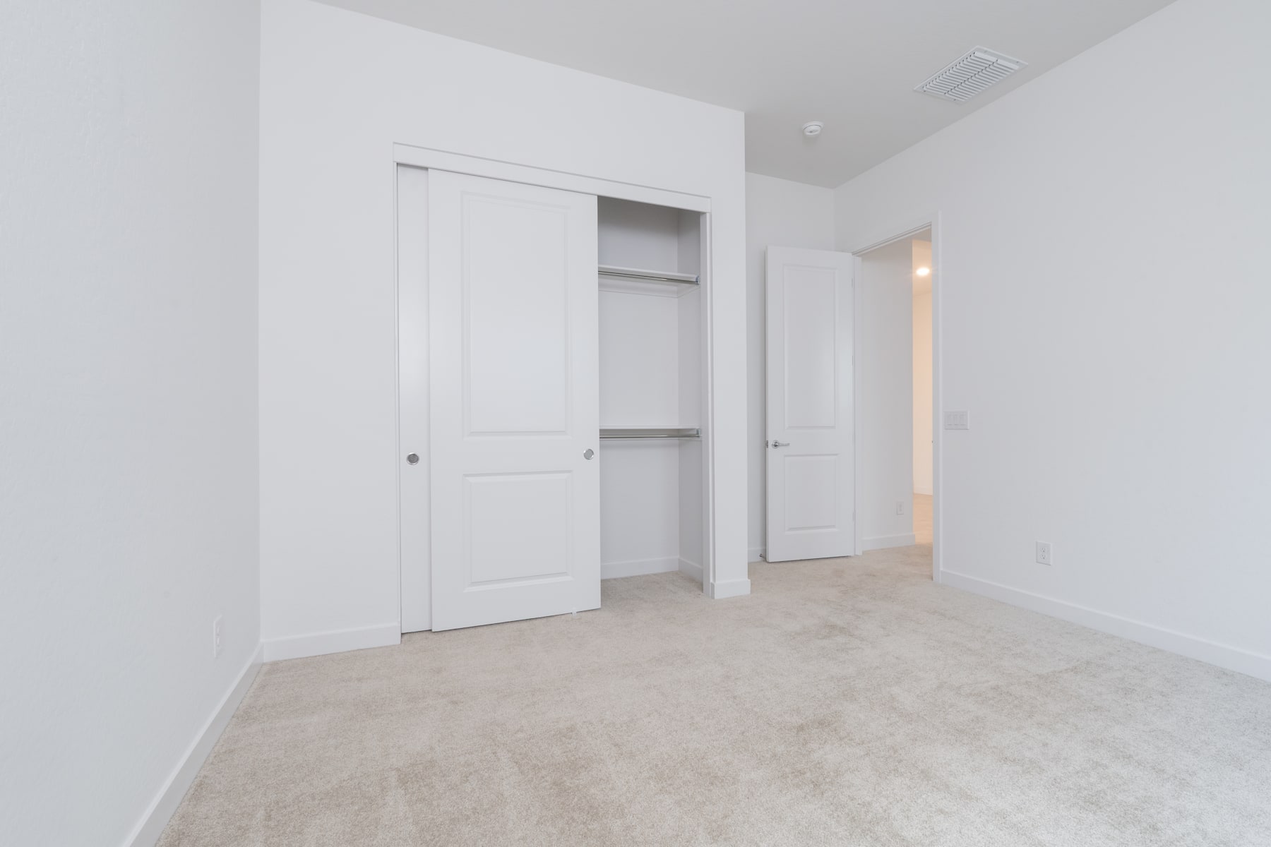 A minimalist, white-walled room with a closet and an open doorway, featuring a light-colored carpeted floor.