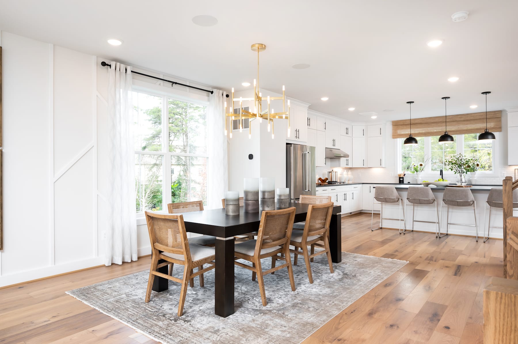 A modern and spacious kitchen and dining area with white walls, wooden floors, and a mix of contemporary and rustic furnishings, including a large dining table, chairs, and a kitchen island with bar stools.