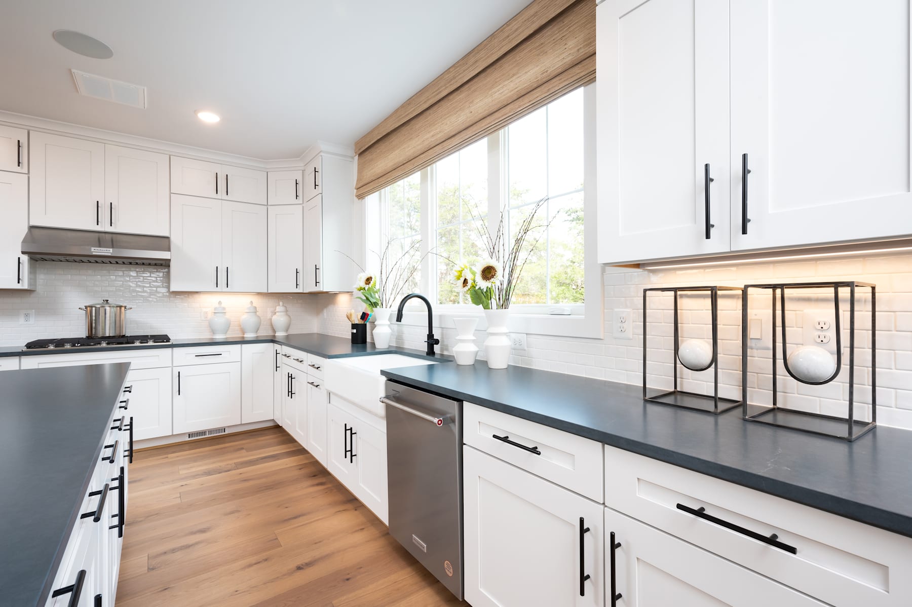 A modern, well-designed kitchen with white cabinets, wooden accents, and a sleek black countertop, featuring a window that provides natural lighting and a view of the outdoors.