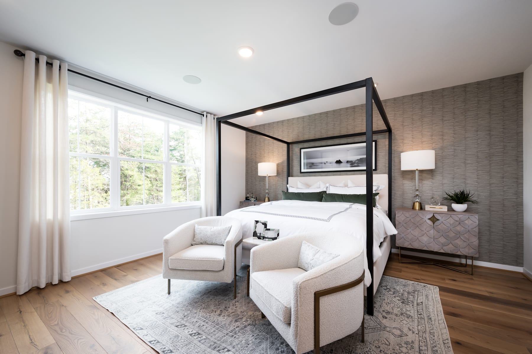 A modern and cozy bedroom with a four-poster bed, a comfortable armchair, and large windows overlooking a lush outdoor scene.