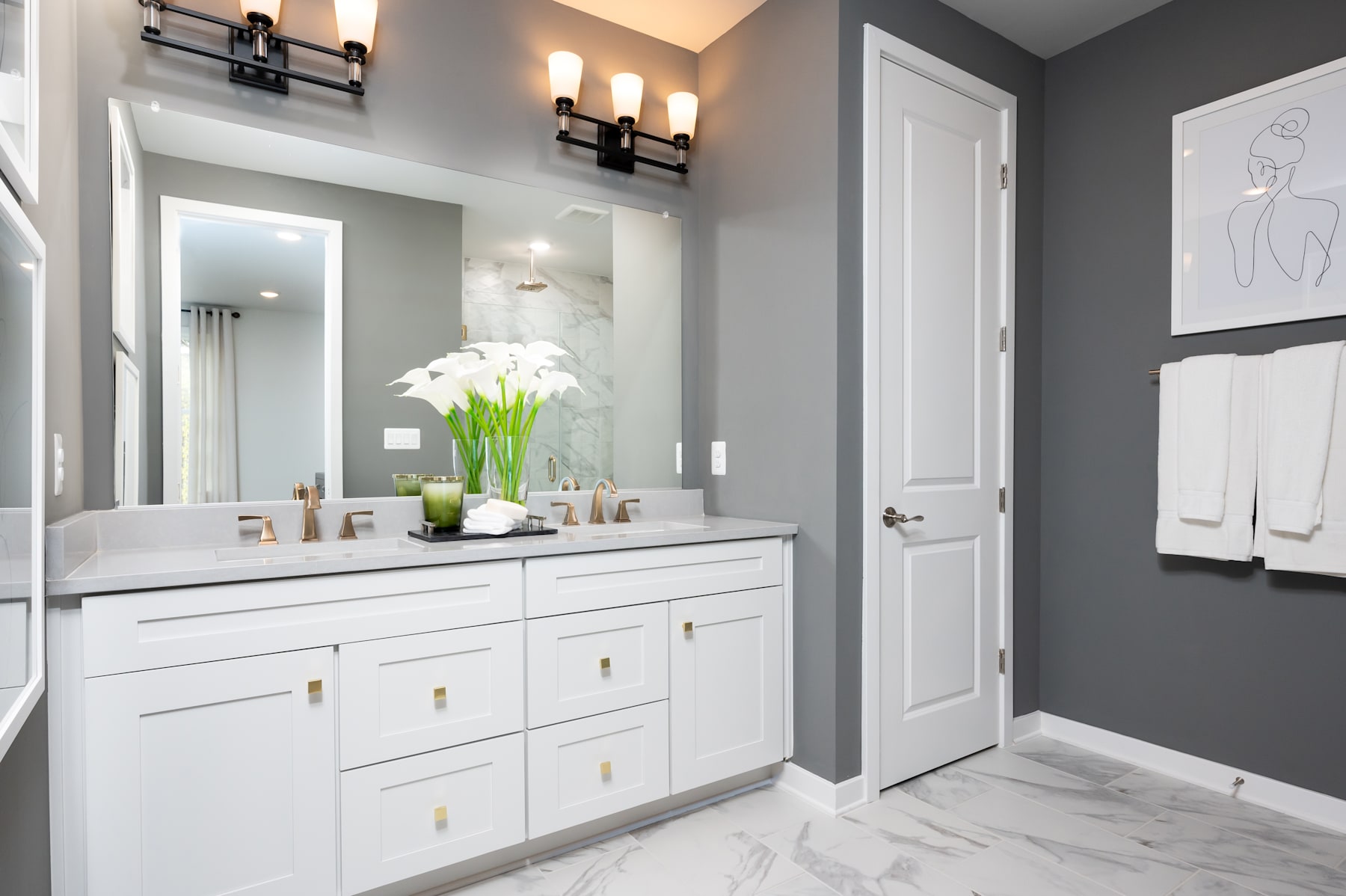 A modern and elegant bathroom with a white vanity, marble flooring, and a vase of white flowers on the counter, set against a gray wall with a framed artwork.