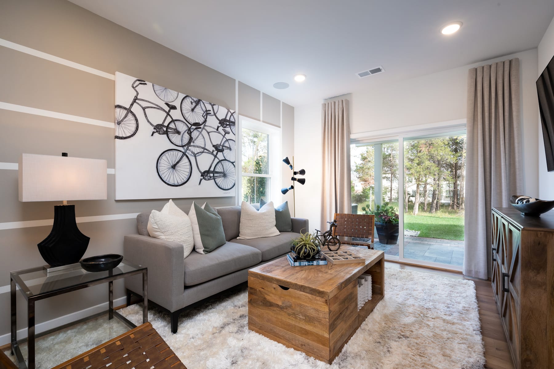 A modern and cozy living room with a gray sofa, a wooden coffee table, and a large wall art featuring bicycles. The room has large windows overlooking a lush outdoor area.