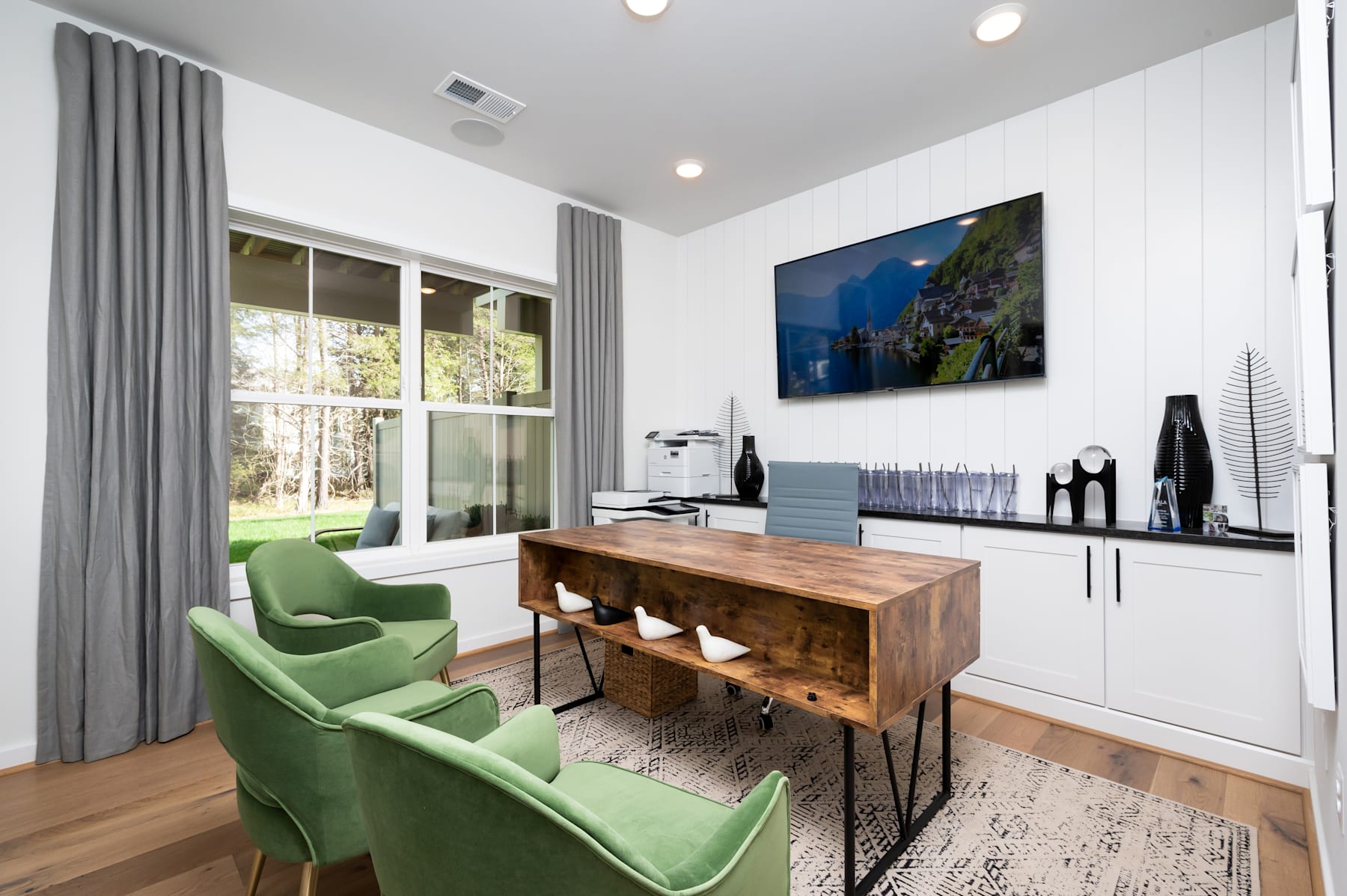 The image depicts a modern and stylish home office space with a large TV mounted on the wall, a wooden console table, and two comfortable green armchairs in the foreground. The room has large windows that provide natural light and a view of the outdoor landscape.