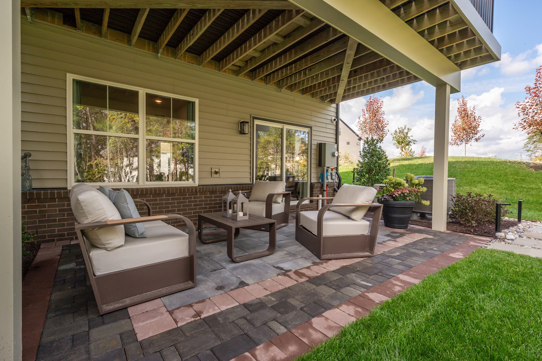 A cozy outdoor seating area with wicker furniture on a brick patio, surrounded by a grassy lawn and a wooden-framed house with large windows.