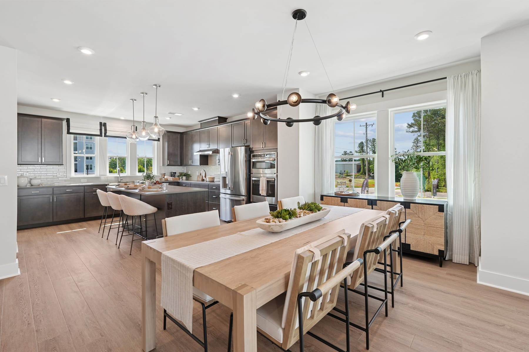 A modern and spacious kitchen with a large wooden dining table, sleek dark cabinets, and a chandelier overhead, surrounded by large windows overlooking a lush outdoor landscape.