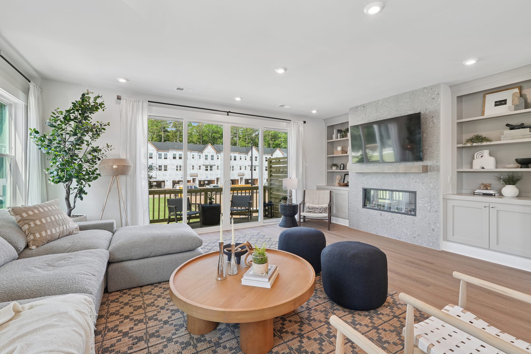 A cozy and modern living room with a large window overlooking a lush outdoor area, featuring a gray sectional sofa, a round wooden coffee table, and various decorative elements.