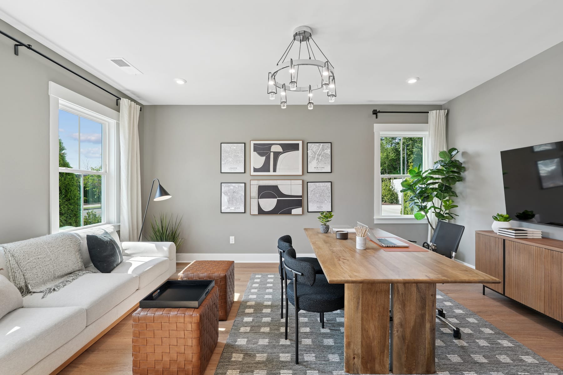 A modern and minimalist living room with a wooden dining table, a gray sofa, and abstract artwork on the walls, complemented by a large window overlooking a lush outdoor scene.
