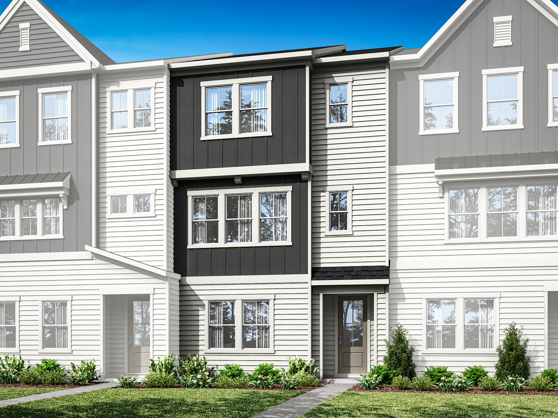 A multi-story residential building with gray siding, white trim, and a blue sky in the background. The building has several windows and a front entrance, surrounded by landscaping with small plants.