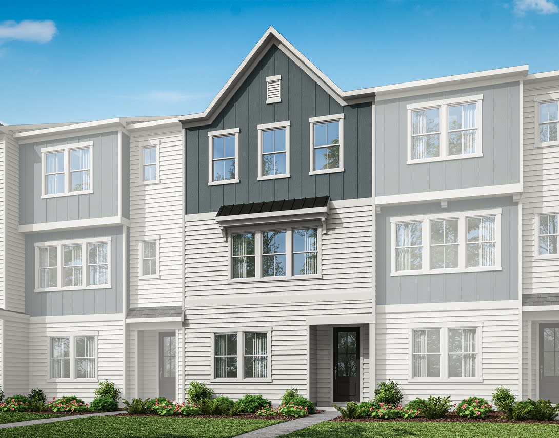 A multi-story townhouse with a gray exterior, white trim, and a gabled roof stands in a well-landscaped setting against a clear blue sky.