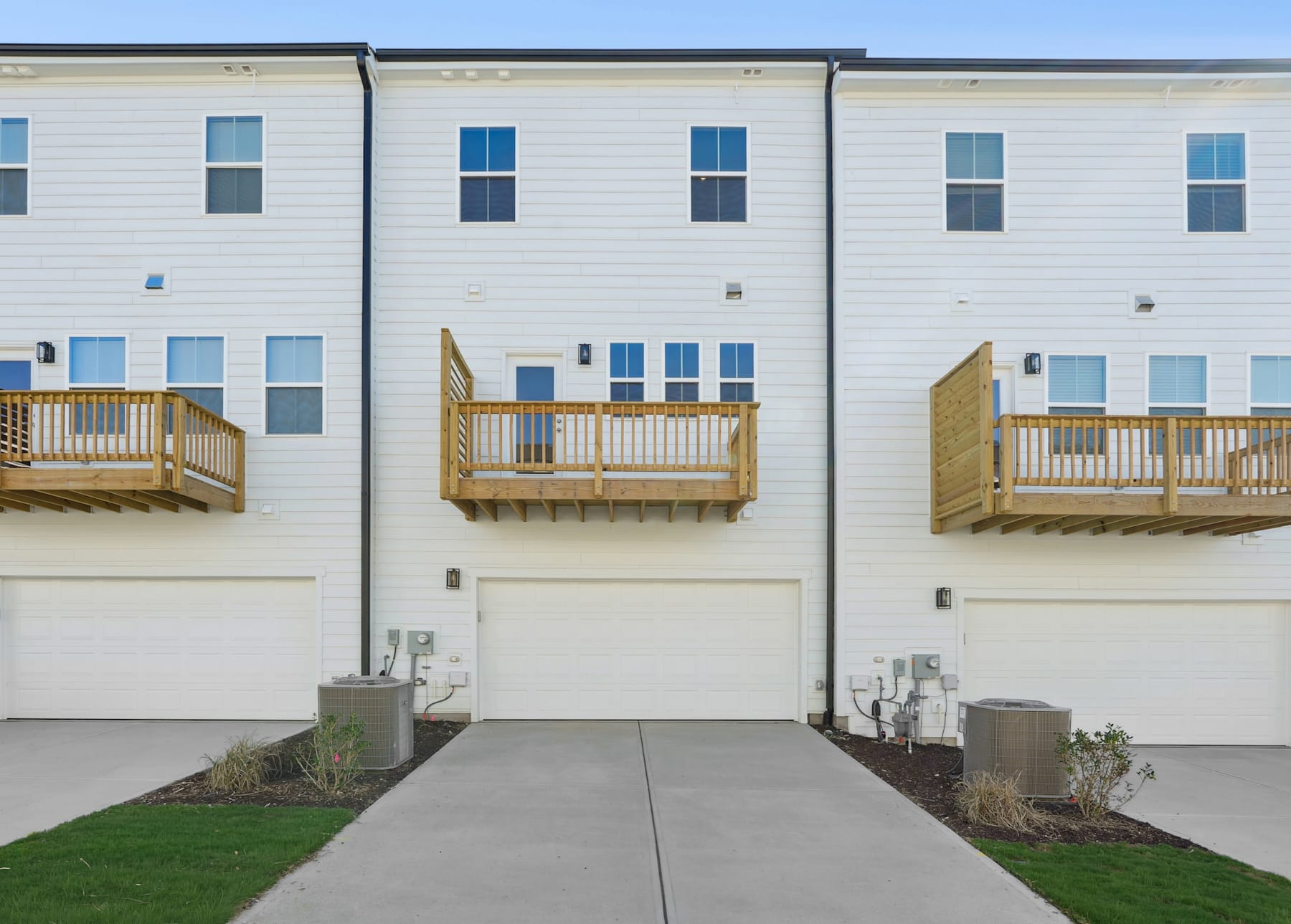 A row of modern, white townhouses with wooden balconies and a paved walkway leading to them, set against a clear blue sky.
