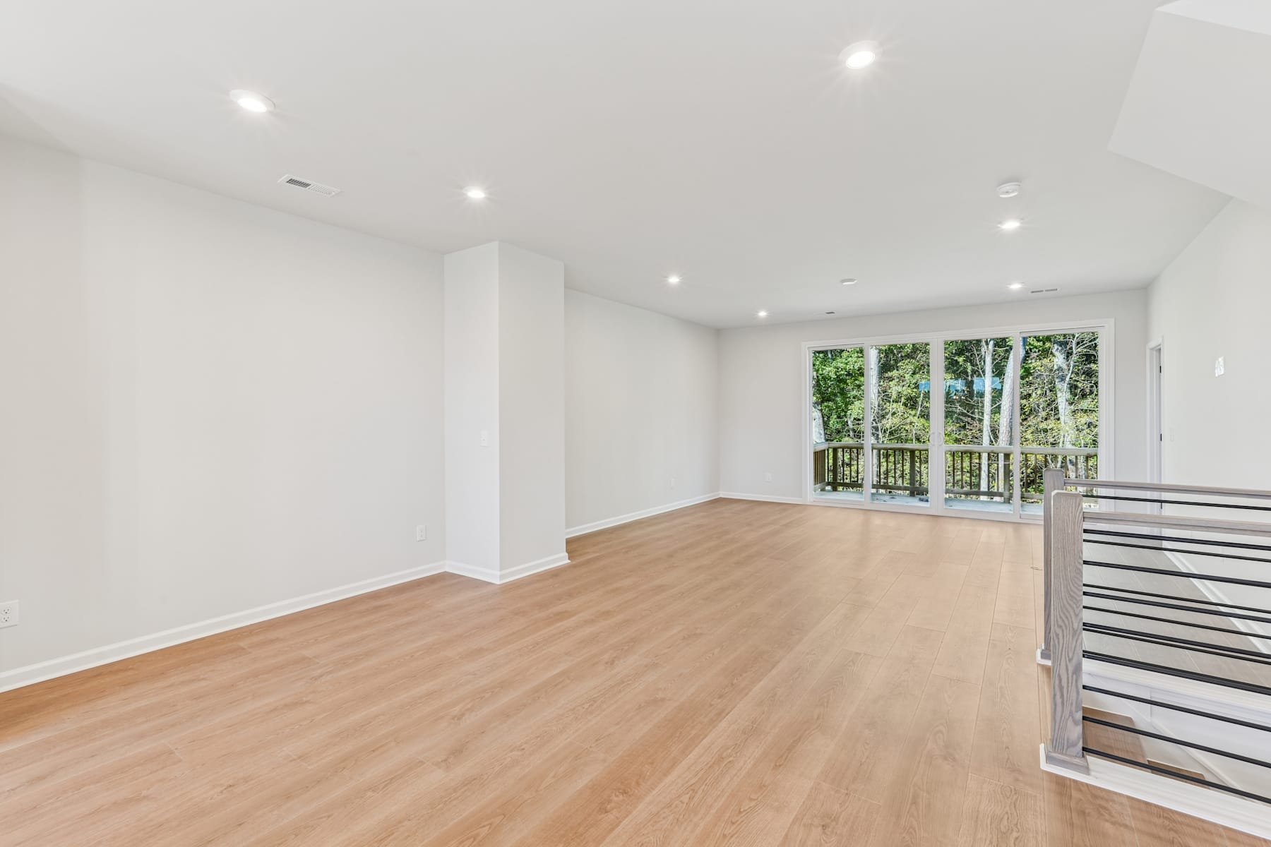 A spacious, well-lit room with wooden flooring, white walls, and large windows overlooking a lush, forested area.