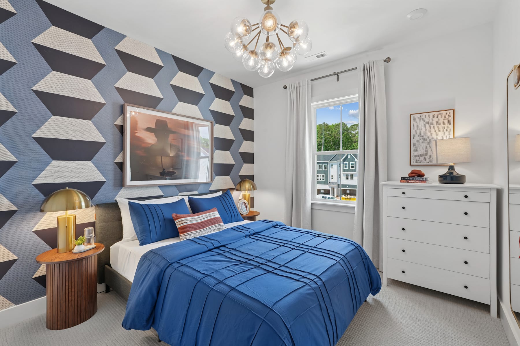 A modern and stylish bedroom with a geometric patterned accent wall, a blue plaid bedspread, and a chandelier overhead, along with a dresser and other furnishings.