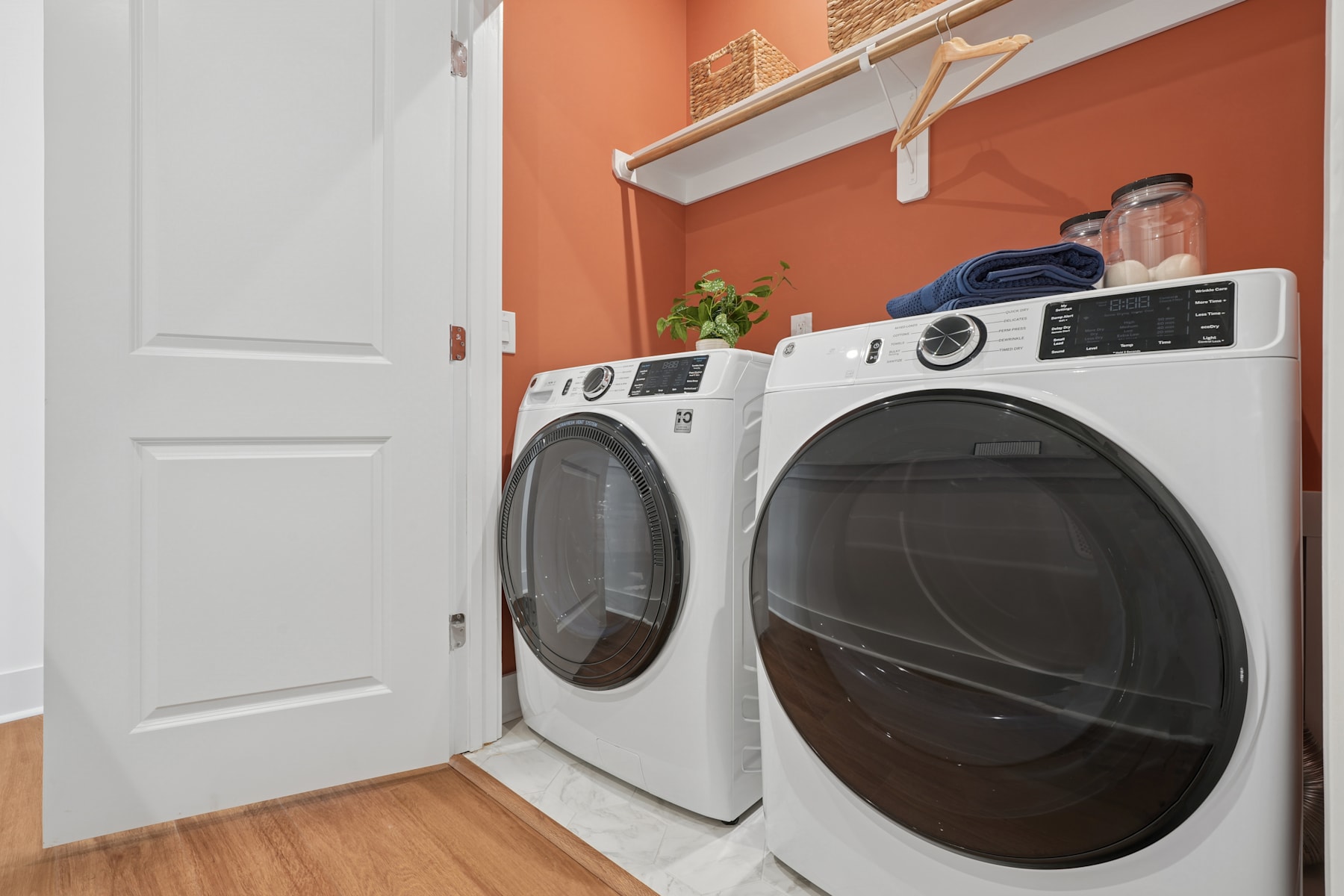 A modern, well-equipped laundry room with a white washer and dryer set against a vibrant orange wall, complemented by a potted plant and other decor elements.