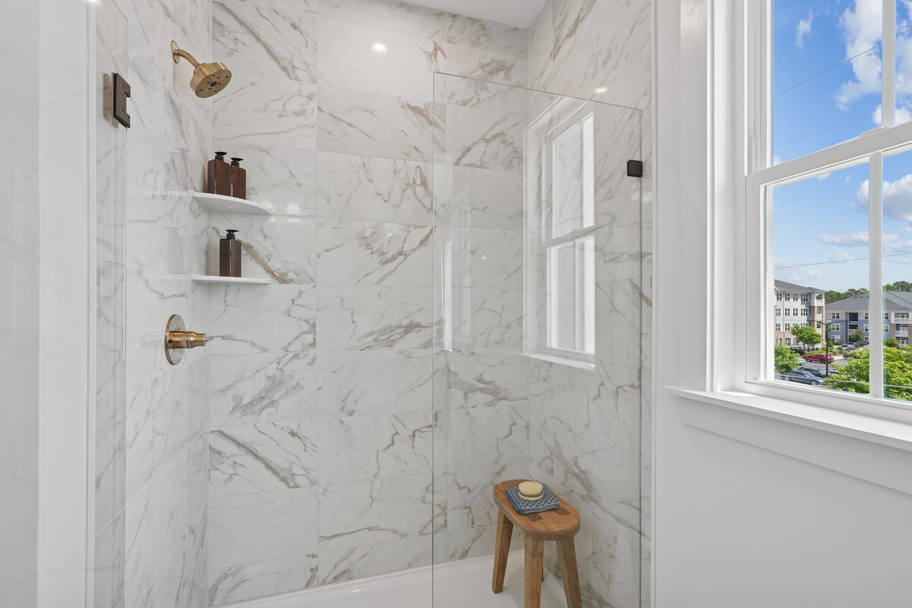 A bright and spacious bathroom with white marble-like walls, shelves displaying various items, and a large window overlooking a scenic outdoor view.
