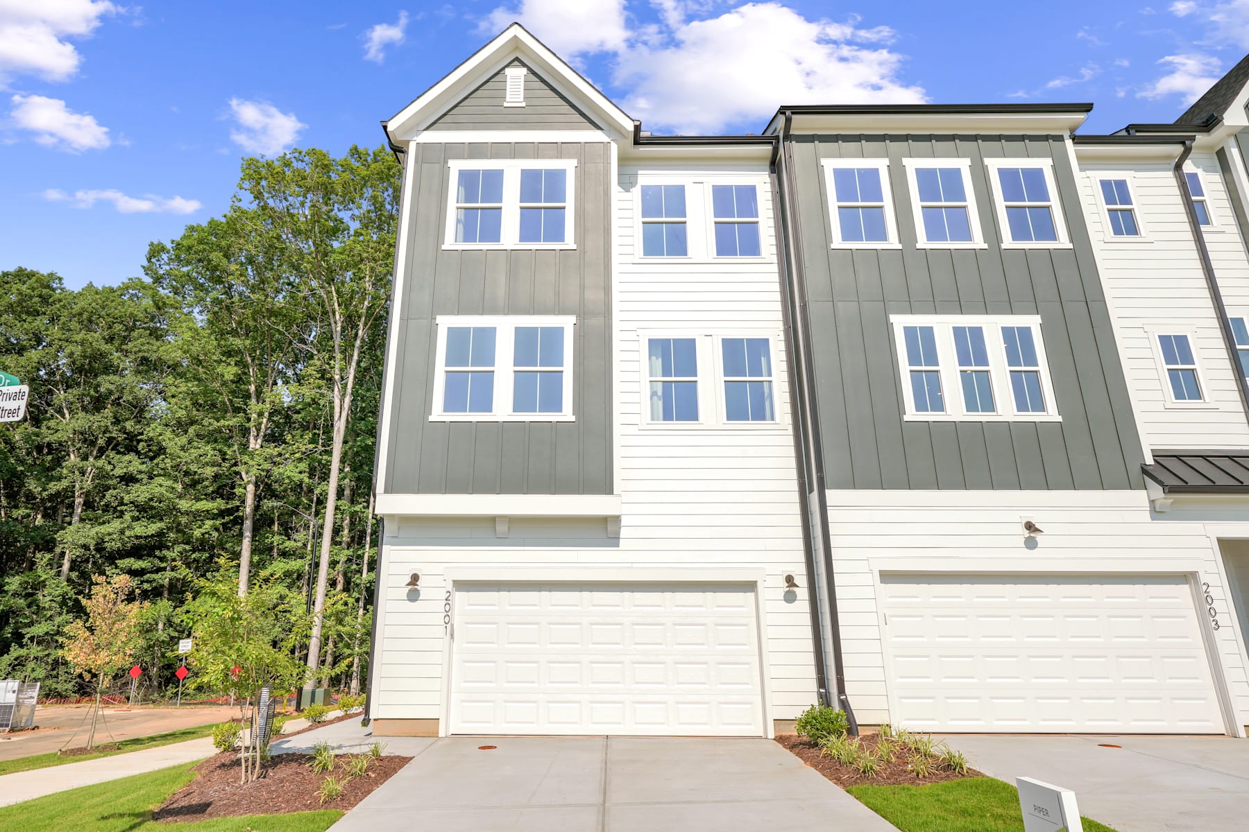 A modern, multi-story townhouse with a garage and a well-landscaped yard, surrounded by lush greenery and a clear blue sky with fluffy clouds.