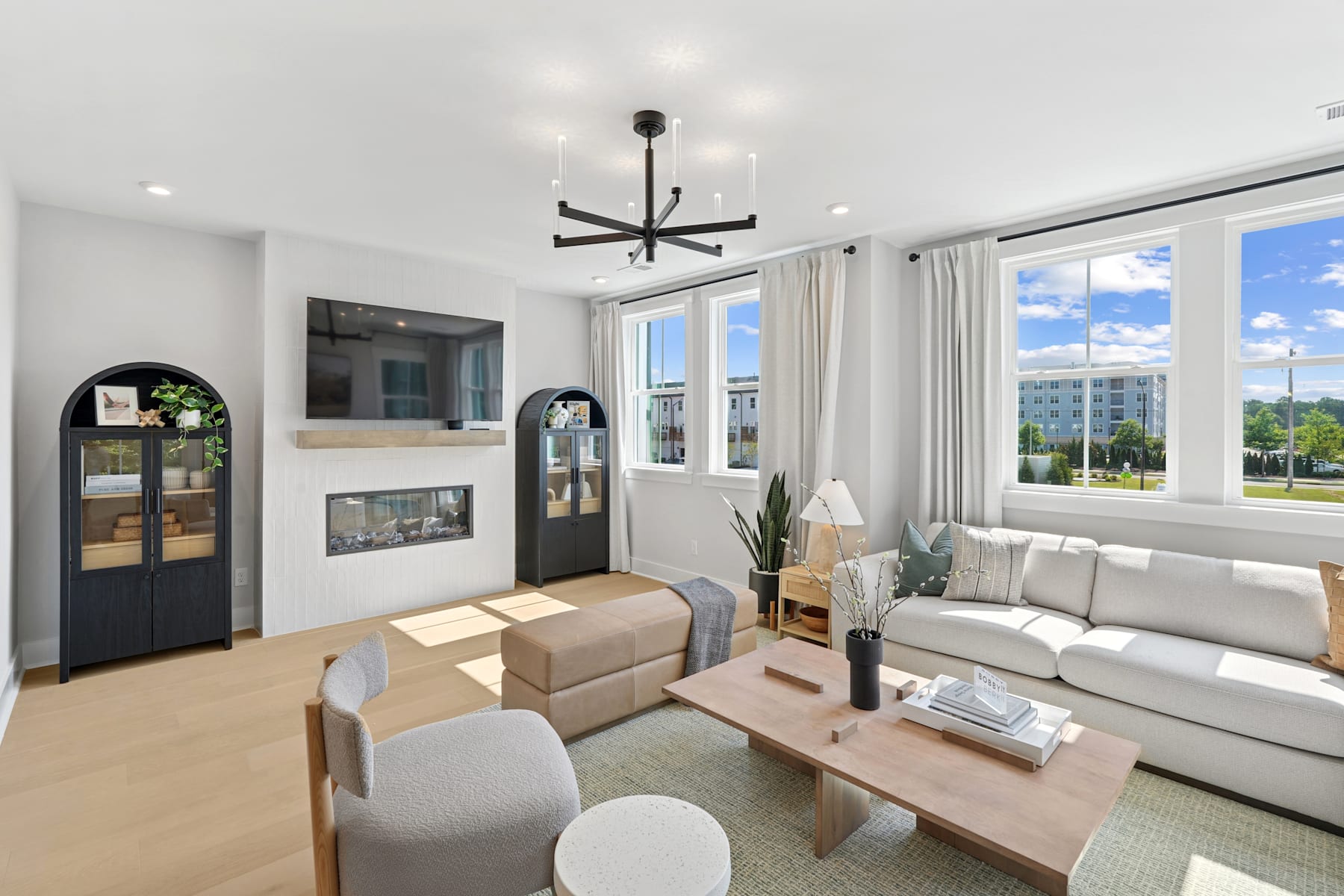 A modern and bright living room with a fireplace, large windows, and a stylish chandelier, surrounded by comfortable furniture and decor.