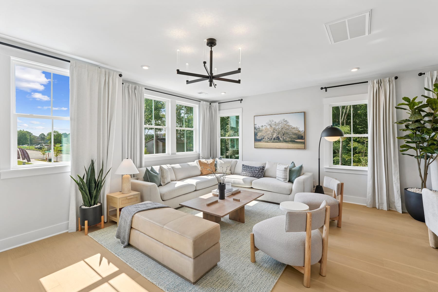 A spacious and well-lit living room with a comfortable sectional sofa, armchairs, and a wooden coffee table, surrounded by large windows that offer a scenic view of the outdoor greenery.