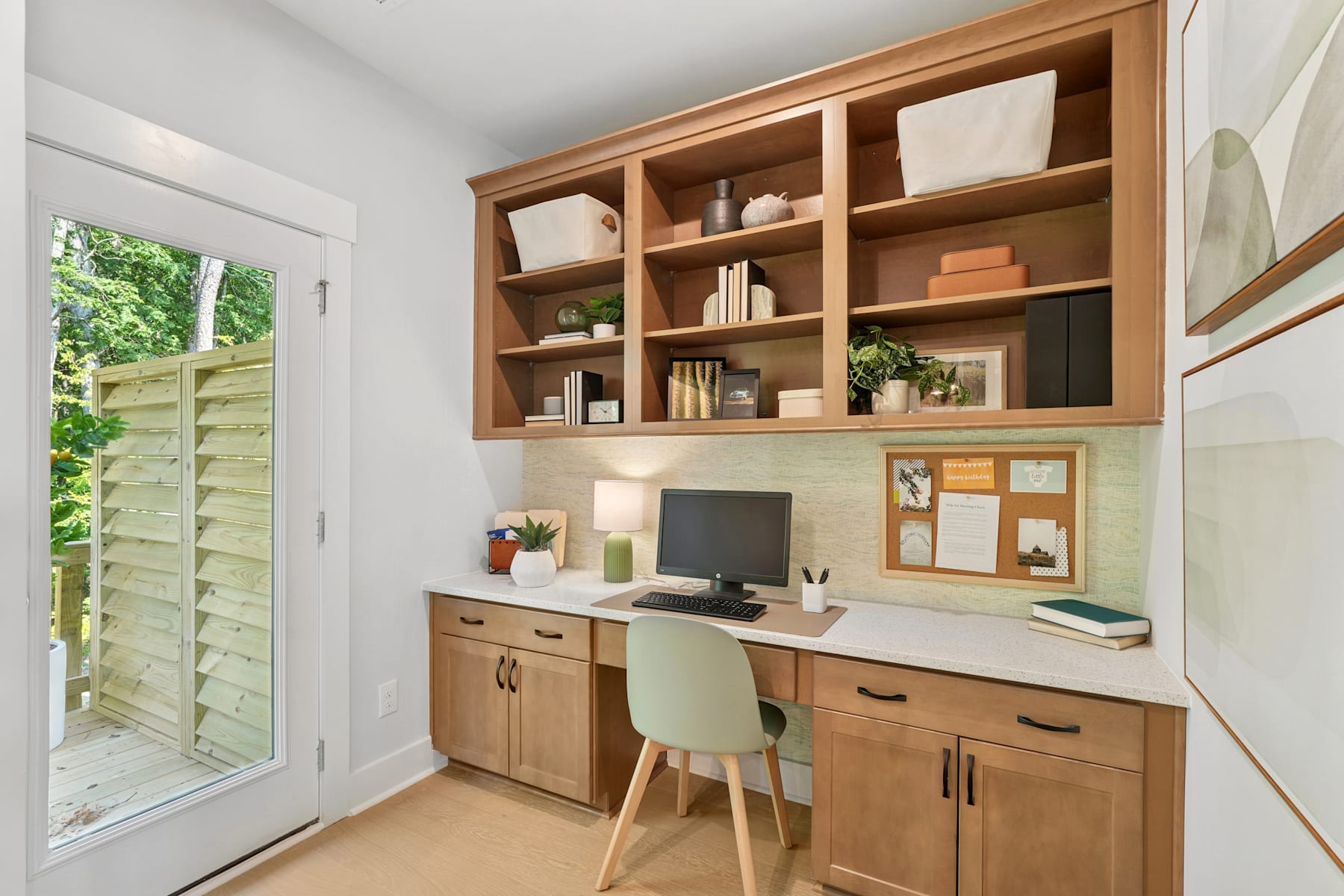 A cozy home office space with wooden shelves, cabinets, and a desk, surrounded by natural light from a window overlooking a lush outdoor area.