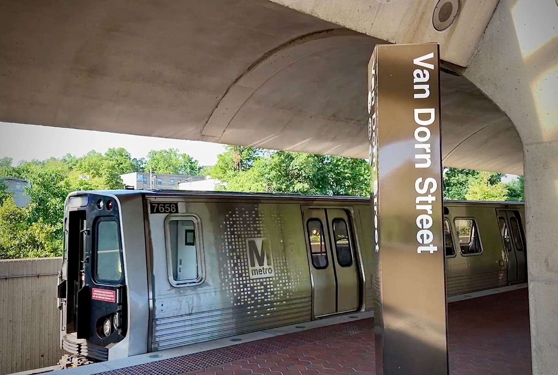 A subway train with the "Van Dorn Street" sign is parked under a concrete overpass, surrounded by greenery in the background.