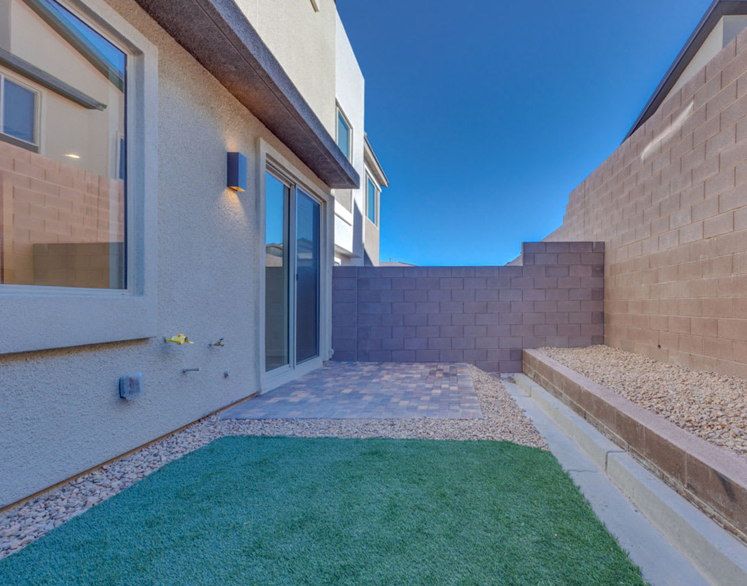 A small, enclosed outdoor space with a grassy area, gravel, and a concrete pathway leading to a modern building with large windows.