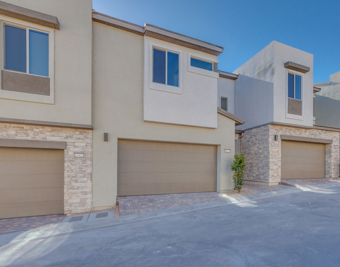 The image shows a modern, multi-unit residential building with a clean, minimalist design featuring a combination of stucco and stone exterior, large windows, and attached garages. The foreground includes a paved driveway or walkway leading to the building.