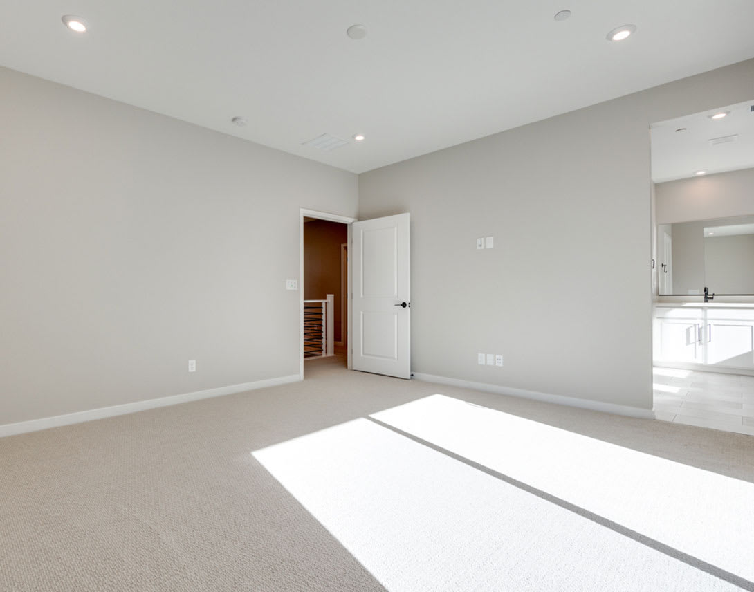 A spacious, well-lit room with a neutral color scheme, featuring a carpeted floor and a doorway leading to another area.