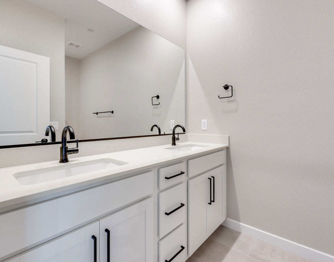 A modern, minimalist bathroom with a white vanity, double sinks, and black hardware against a plain white wall background.