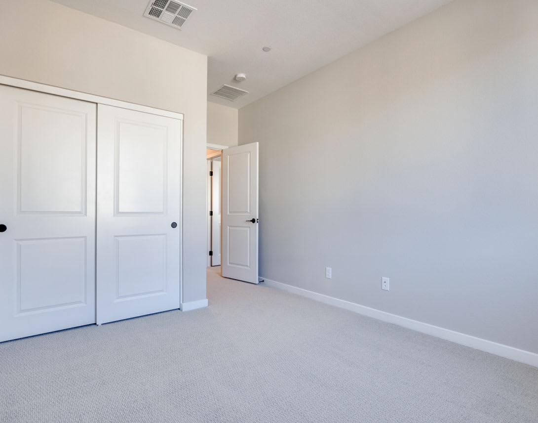 A spacious, minimalist bedroom with white walls, sliding closet doors, and a carpeted floor.