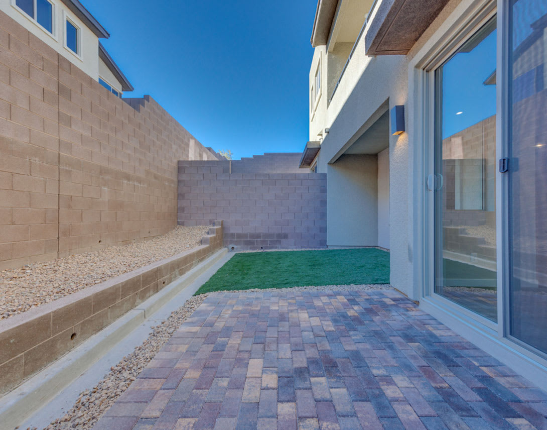 A paved walkway leads to a small grassy area surrounded by a modern building with large windows, set against a clear blue sky.