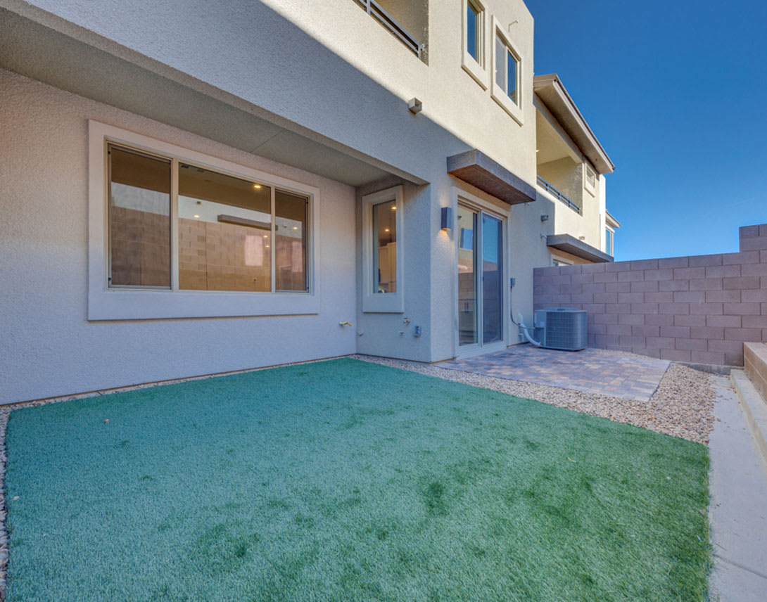 A modern, two-story residential building with a small yard in the foreground, featuring a grassy area and a paved walkway leading to the entrance.