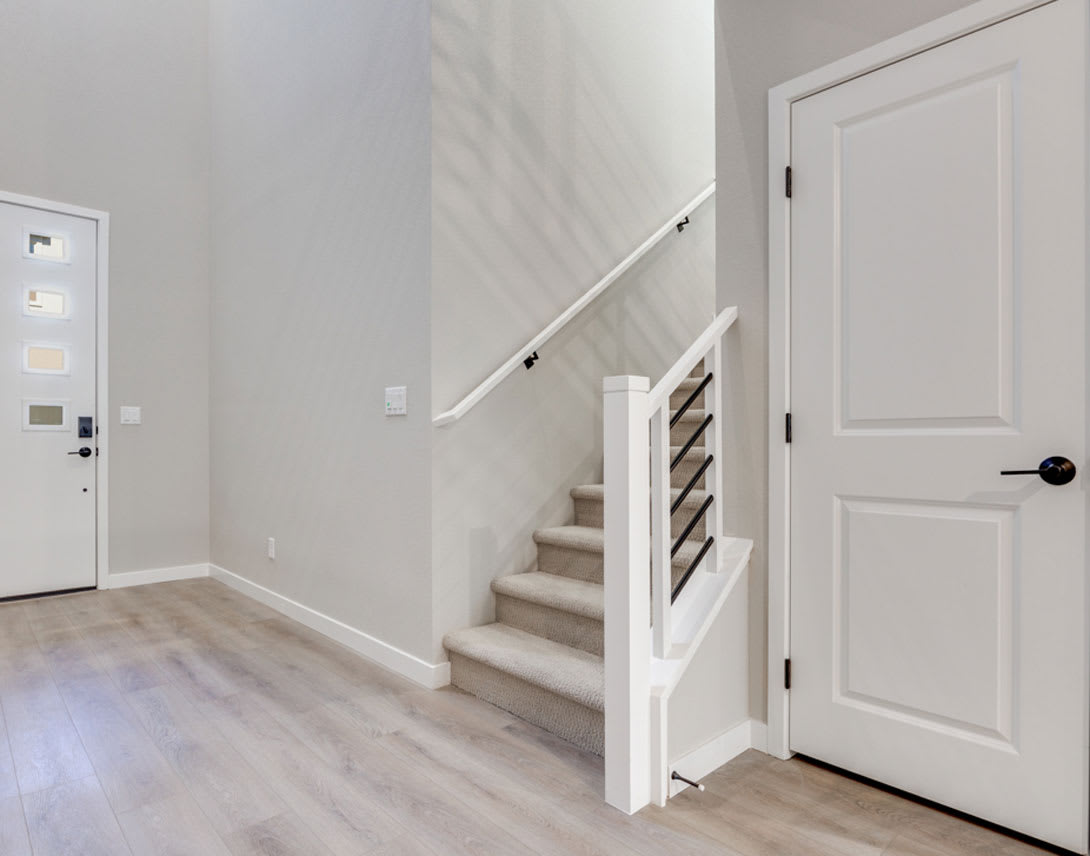 A bright and airy entryway with a white staircase leading up, wooden floors, and a white door in the background.