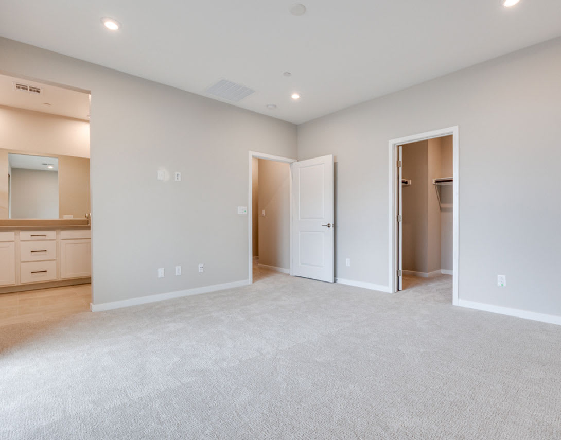 A spacious, well-lit room with a neutral color scheme, featuring a carpeted floor, a vanity area, and multiple doors leading to other parts of the space.